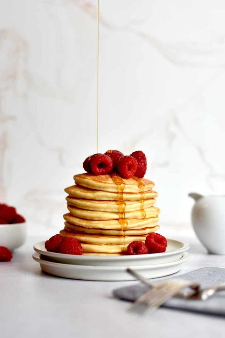 Stack of sweet cream pancakes on a white plate garnished with fresh raspberries and a drizzle of maple syrup pouring down.