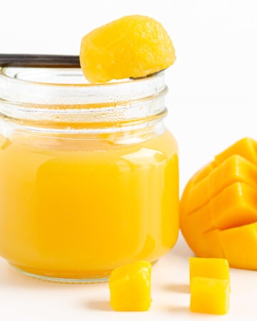 A spoonful of mango jelly set across the top of a jar of mango jelly next to some fresh cut mango.