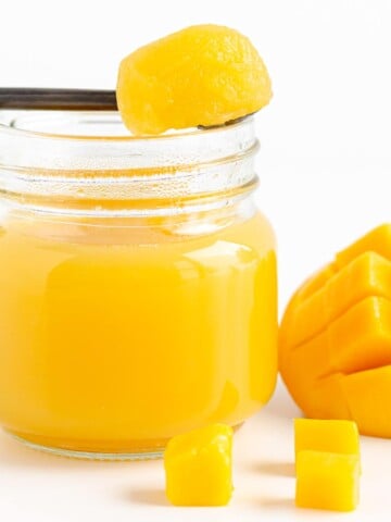 A spoonful of mango jelly set across the top of a jar of mango jelly next to some fresh cut mango.