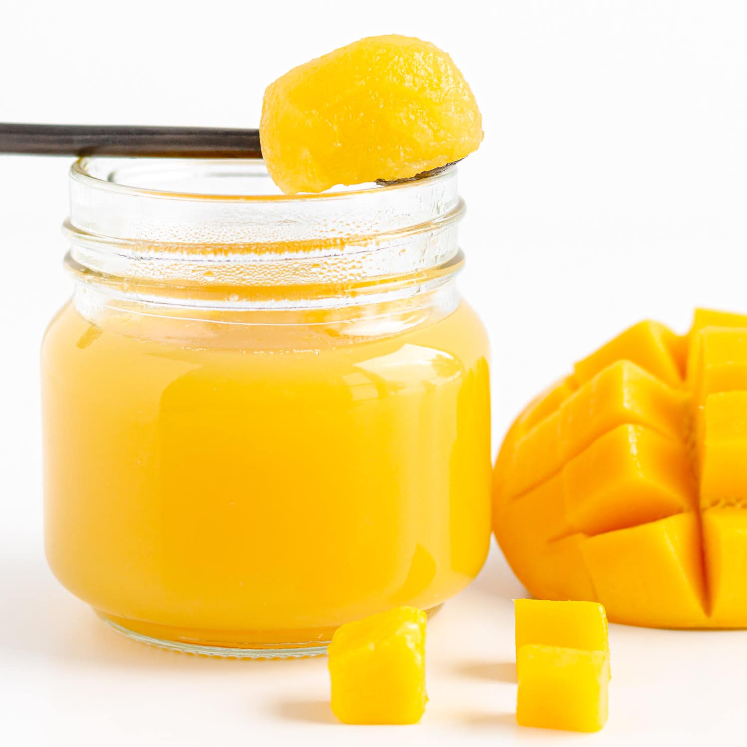 A spoonful of mango jelly set across the top of a jar of mango jelly next to some fresh cut mango.