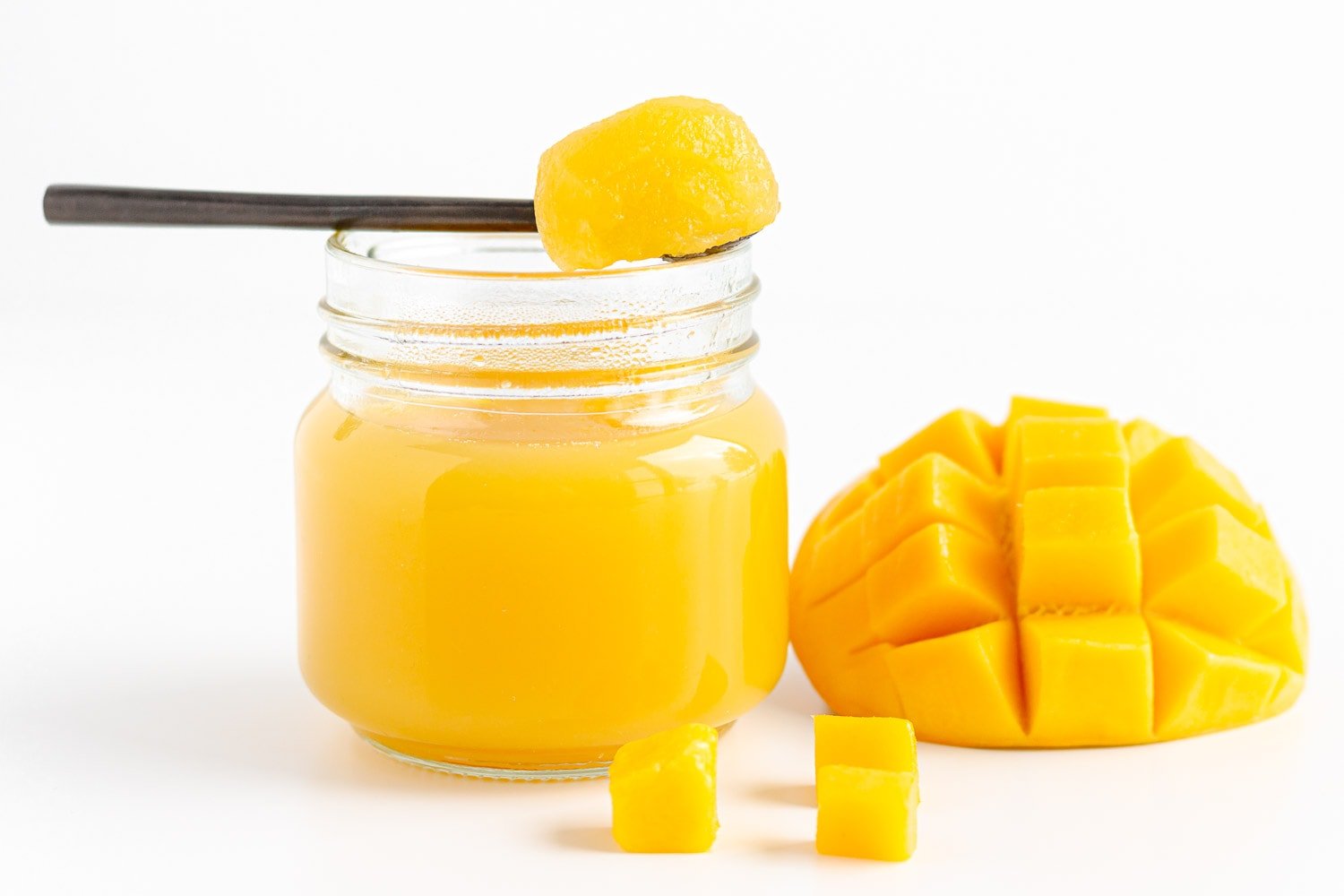 Mango jelly on a spoon set over a small glass jar filled with mango jelly beside some cut up fresh mango.