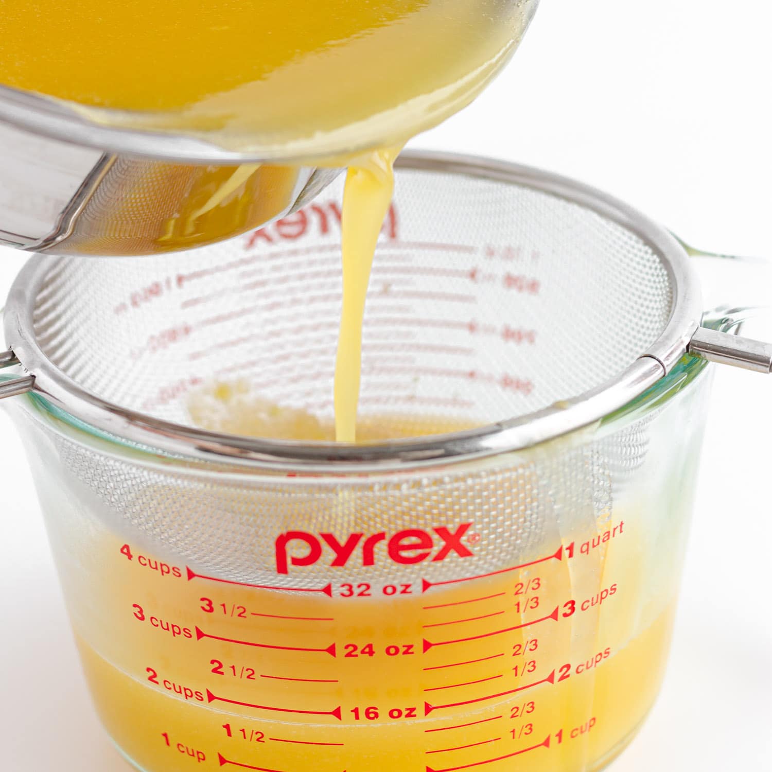 Pouring mango jelly mixture from the saucepan through a fine mesh sieve set over a large glass measuring cup.