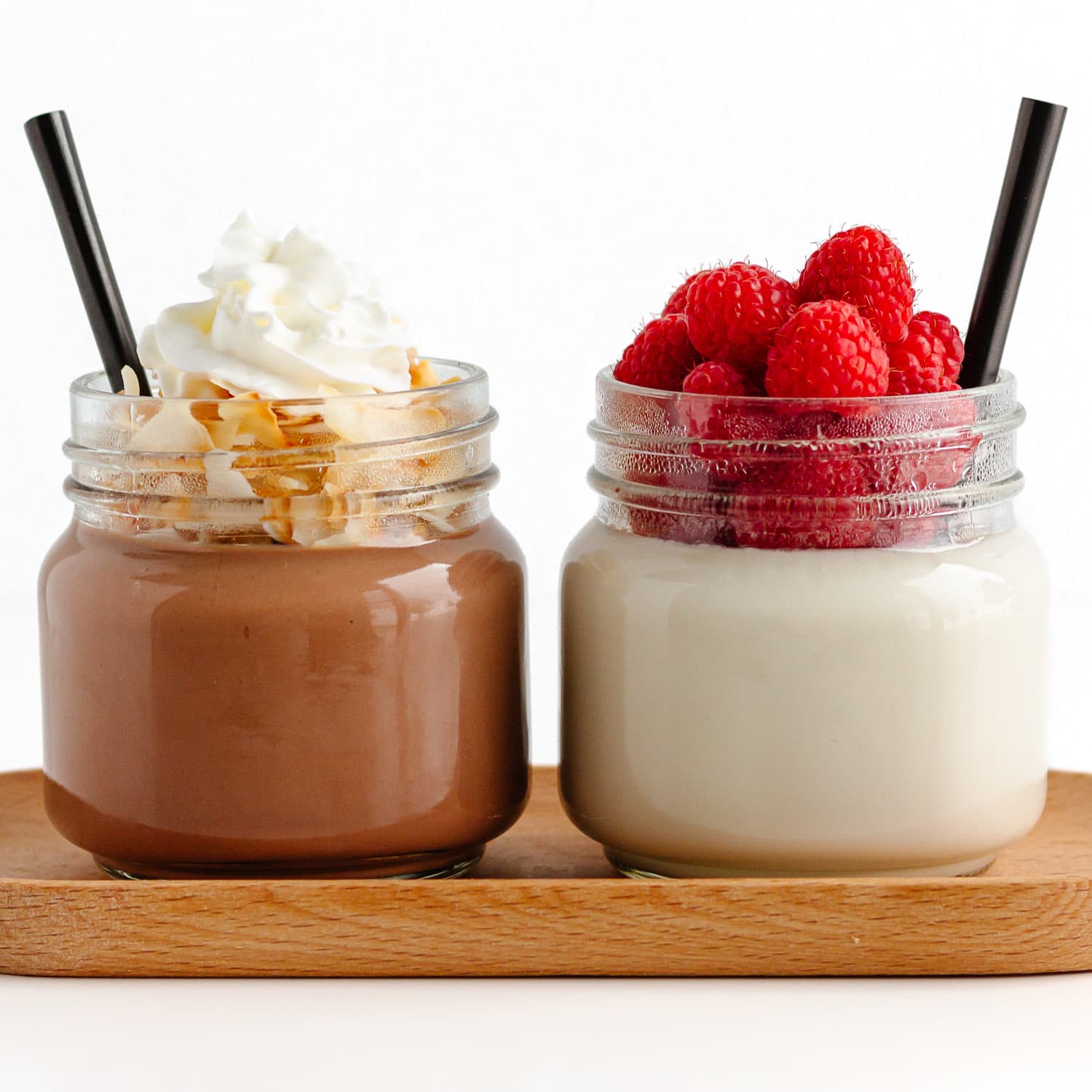 Two glass jars of coconut jello - one chocolate flavor topped with coconut flakes and whipped cream and one vanilla flavor topped with raspberries.