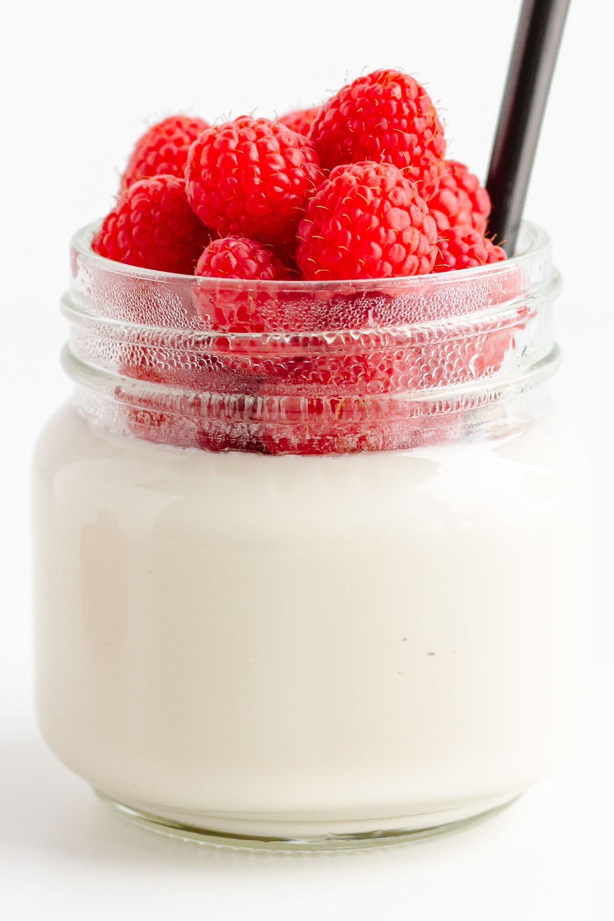 Glass jar of vanilla coconut milk jelly topped with raspberries and spoon sticking out.