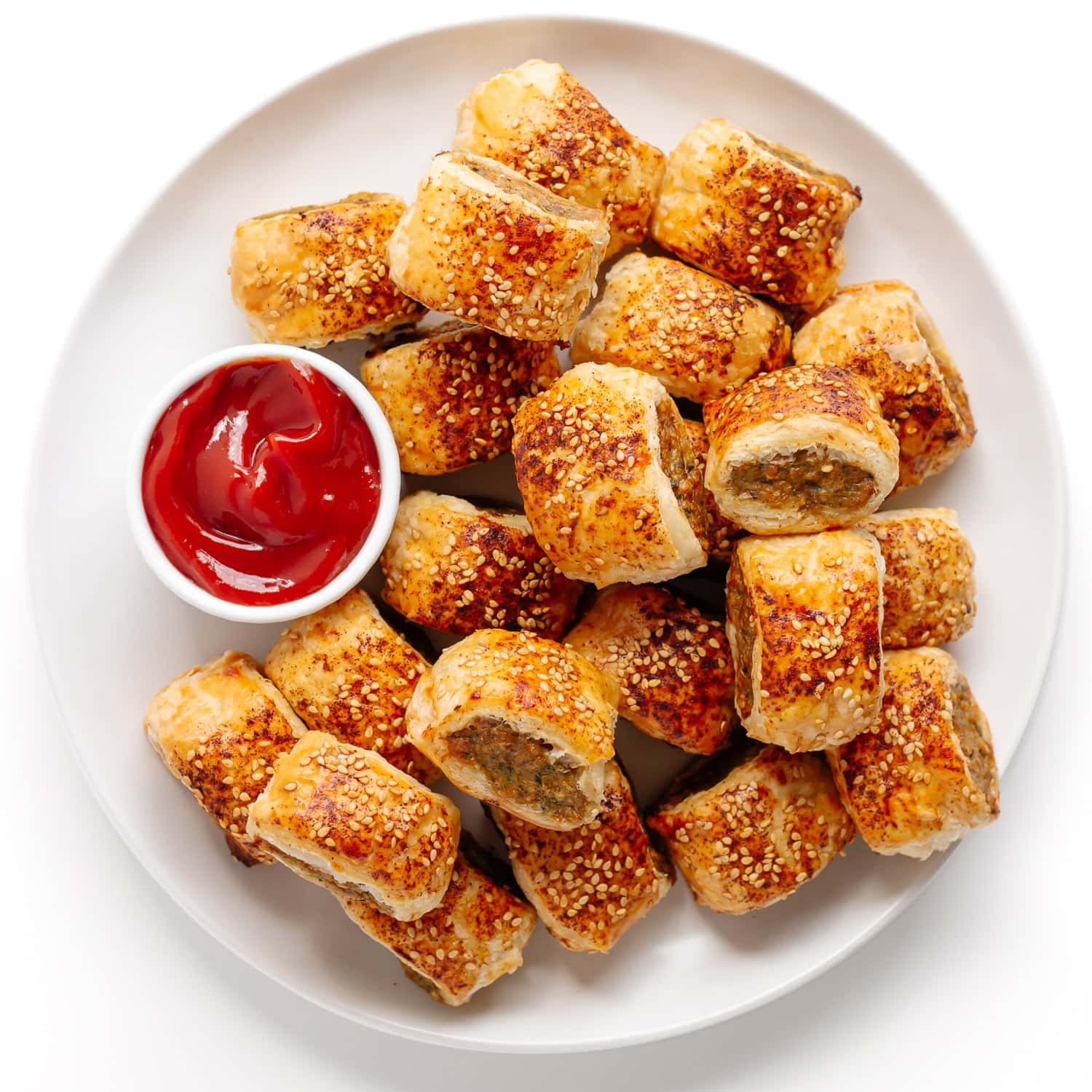 Homemade chicken sausage rolls piled on a large white plate with small bowl of ketchup.