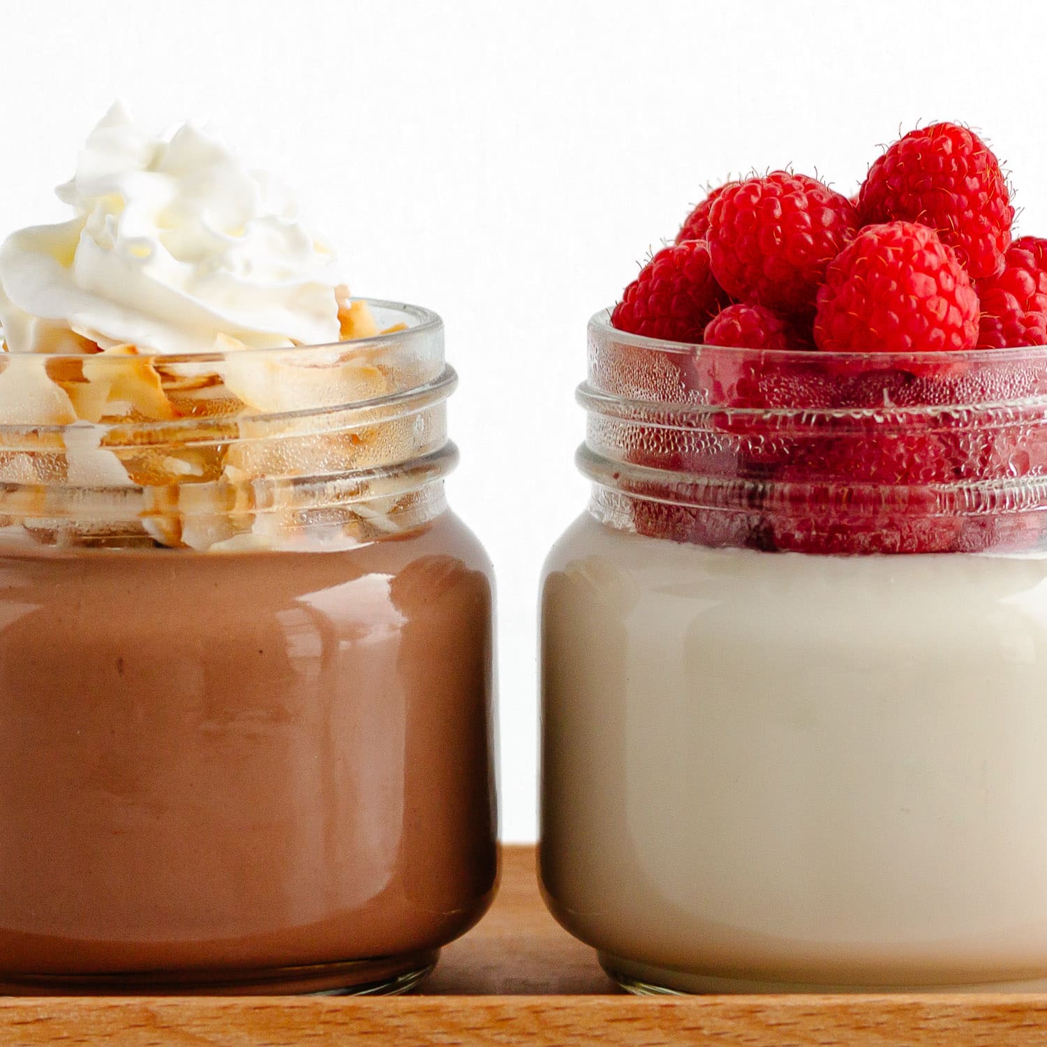 Closeup of two jars coconut jelly, one chocolate jelly topped with coconut flakes and whipped cream and one vanilla coconut jelly topped with fresh raspberries.