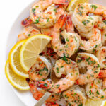 Garlic ginger pan seared shrimp on a white serving plate garnished with lemon slices and chopped parsley.