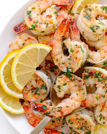 Garlic ginger pan seared shrimp on a white serving plate garnished with lemon slices and chopped parsley.