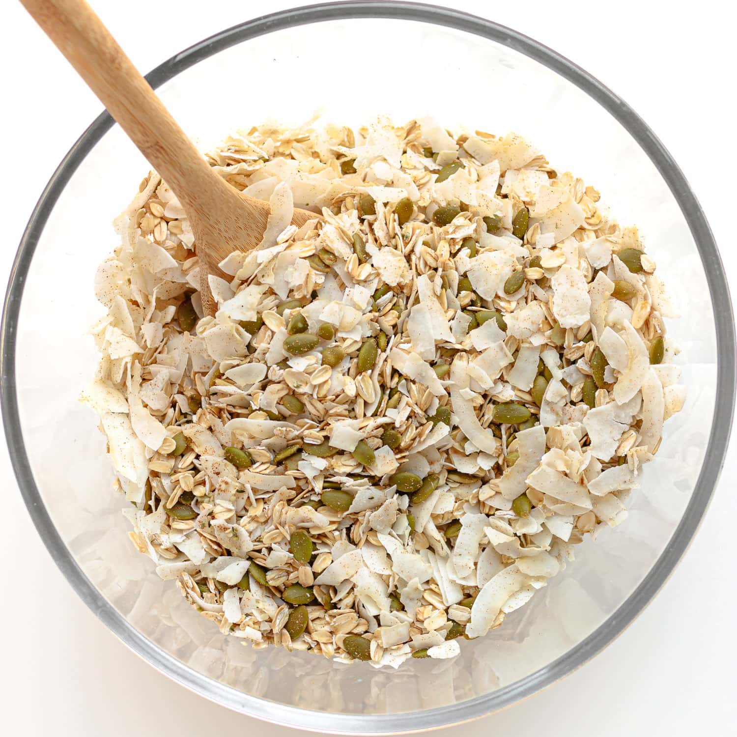 Dry ingredients for coconut granola being mixed together with a wooden spoon in a large glass bowl.