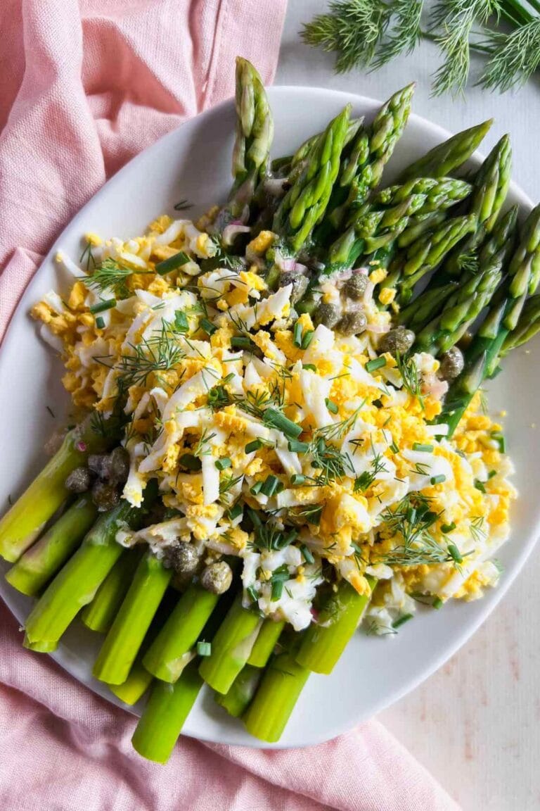 Cooked asparagus on a serving platter topped with grated egg, caper vinaigrette and fresh herbs.