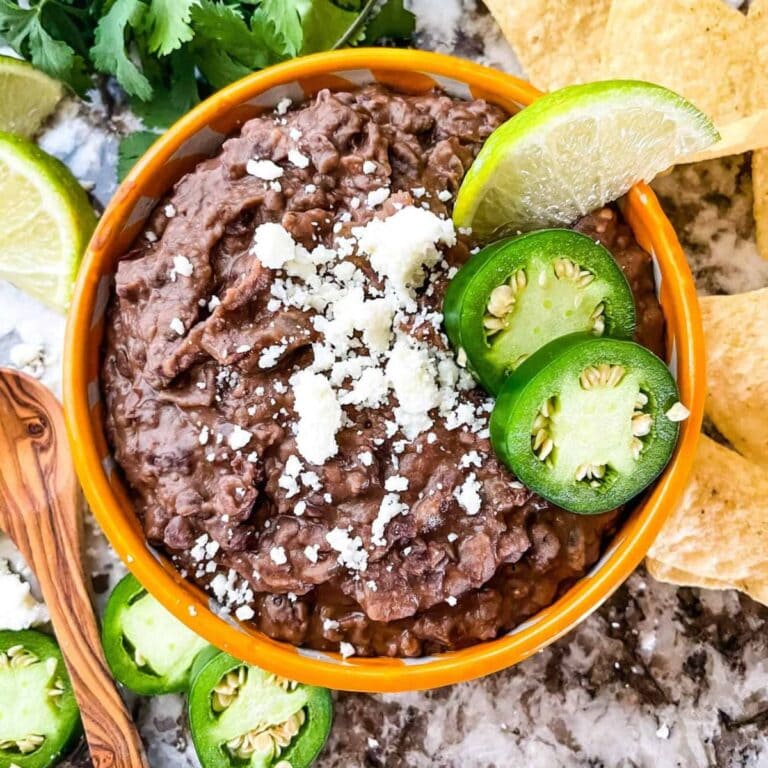 Bowl of black refried beans garnished with jalapeno slices, lime wedge and crumbled cotija cheese.