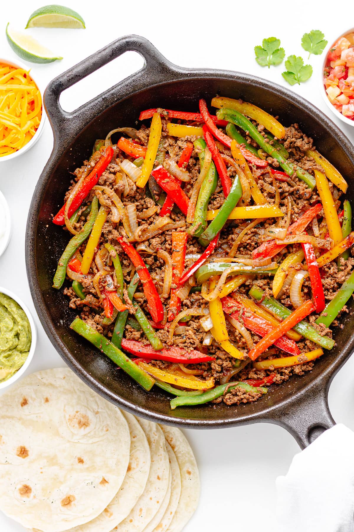 Cast iron skillet of ground beef fajitas mixture surrounded by tortillas and various toppings.