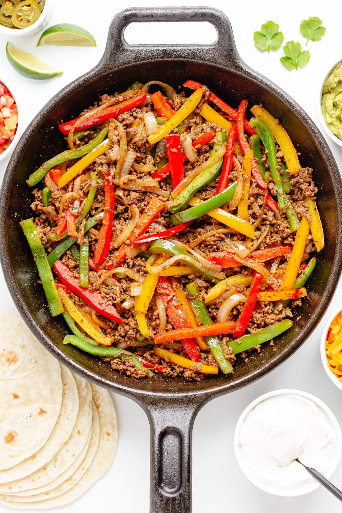Ground beef and fajita veggie mixture in a cast iron skillet surrounded by tortillas and a variety of fajita toppings.