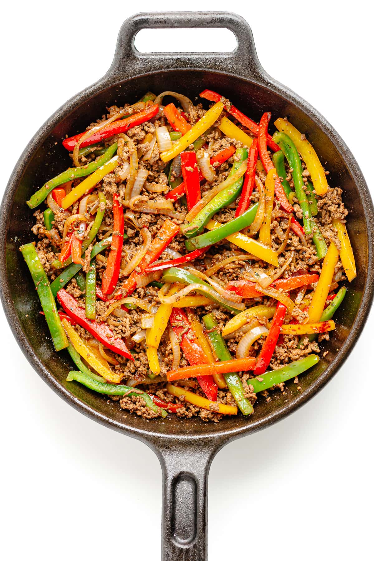 Seasoned ground beef and fajita veggies in a cast iron skillet on a white background.