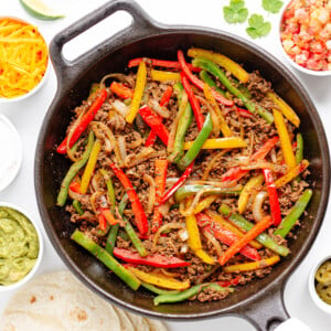 Ground beef fajitas mixture in a cast iron skillet surrounded by a variety of fajita toppings and tortillas.