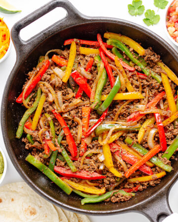 Ground beef fajitas mixture in a cast iron skillet surrounded by a variety of fajita toppings and tortillas.