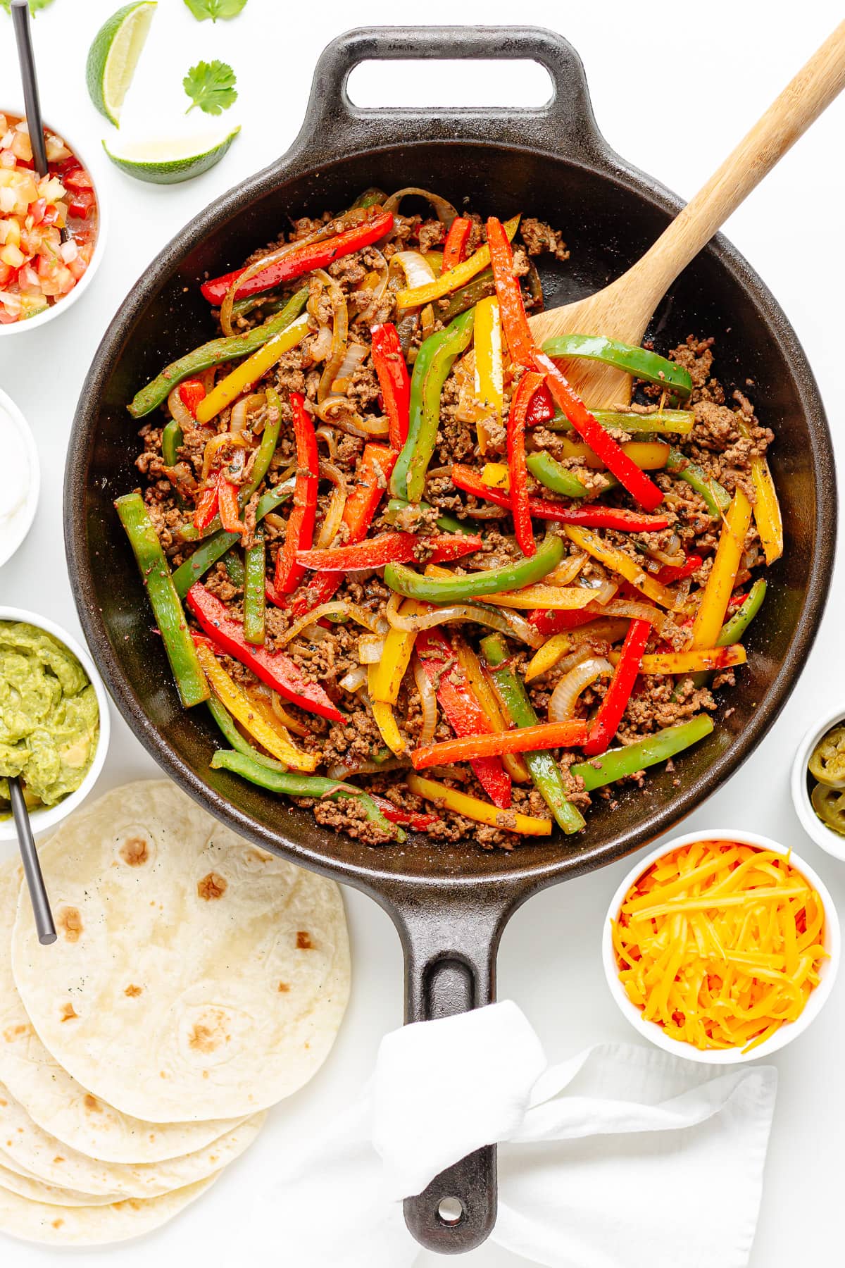 Ground beef fajitas filling in a cast iron skillet with wooden spoon, surrounded by a variety of toppings and tortillas.