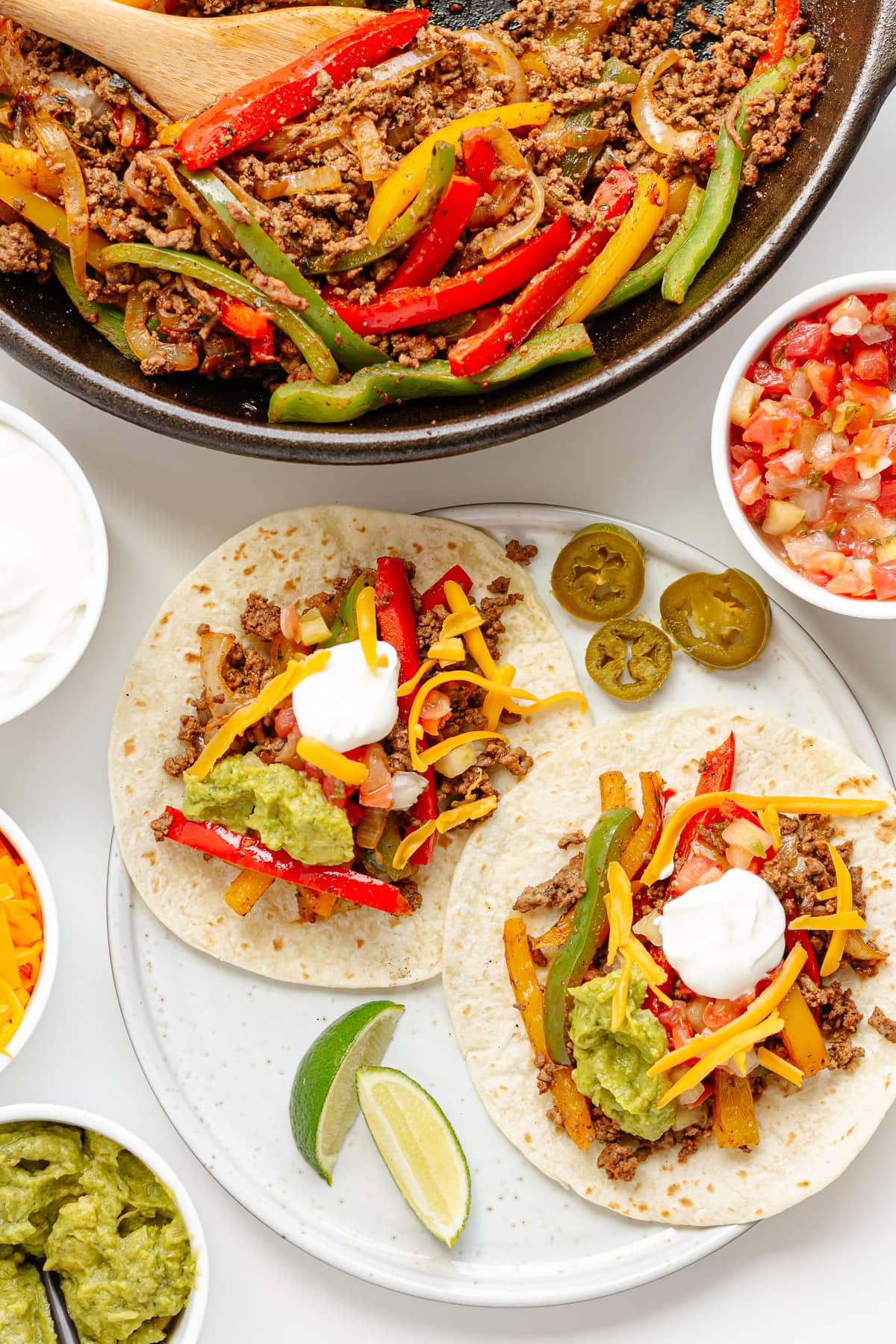 Two ground beef fajitas on flour tortillas on a plate with toppings and skillet of fajita mixture next to it.