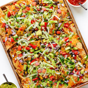 Loaded veggie nachos on a sheet pan with two small bowls of salsa and guacamole beside it.