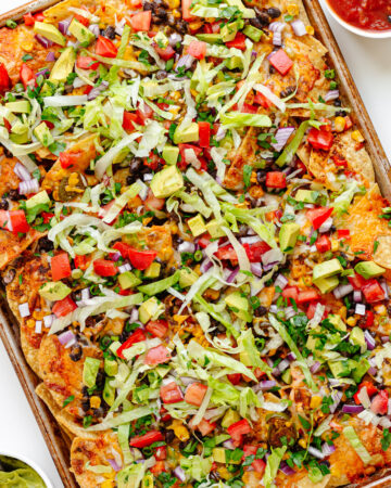 Loaded veggie nachos on a sheet pan with two small bowls of salsa and guacamole beside it.