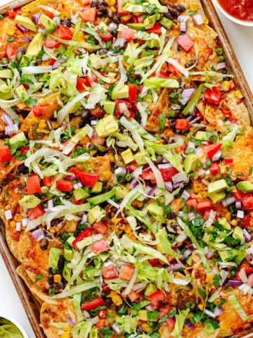 Loaded veggie nachos on a sheet pan with two small bowls of salsa and guacamole beside it.