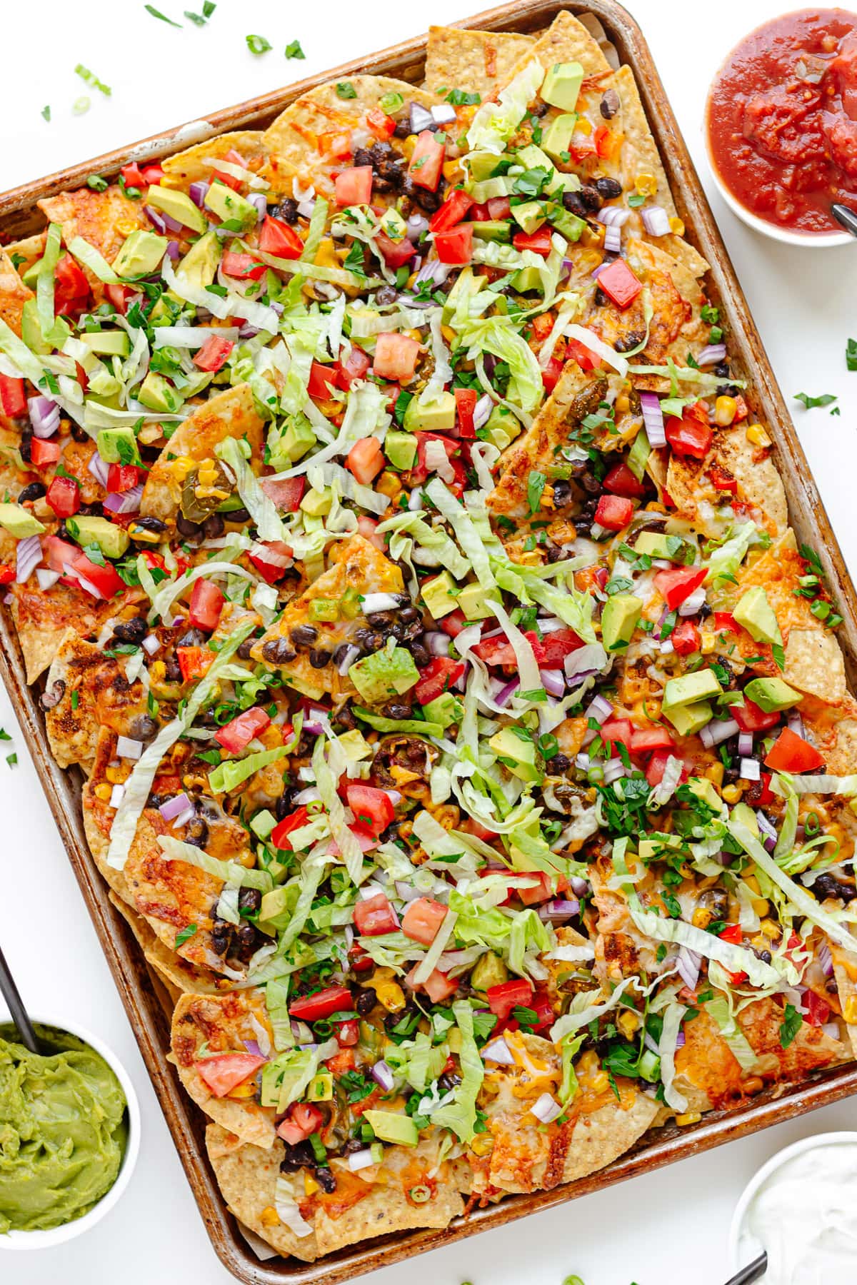 Loaded veggie nachos on a sheet pan with bowls of salsa, guacamole and sour cream around it.