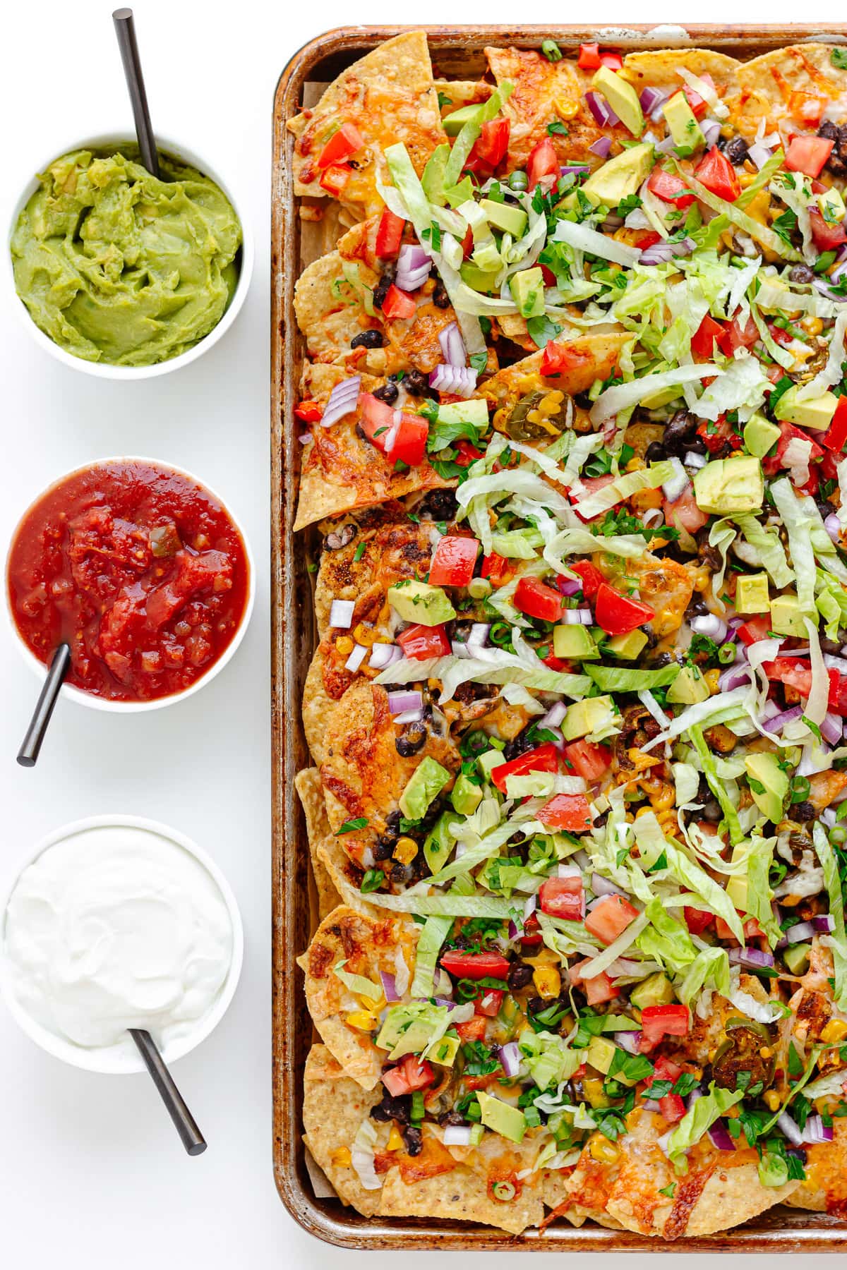 Loaded veggie sheet pan nachos with small bowls of guacamole, salsa and sour cream lined up beside it.