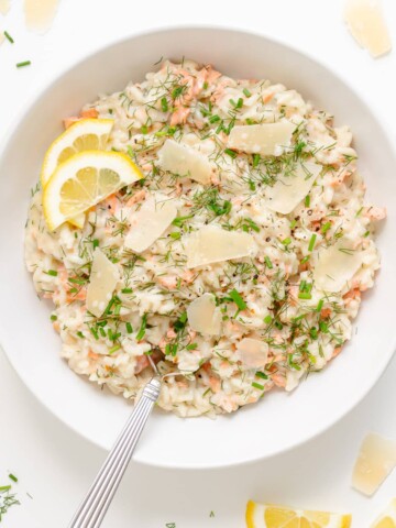 White bowl of salmon risotto garnished with lemon slices, shaved parmesan, fresh dill and chives.