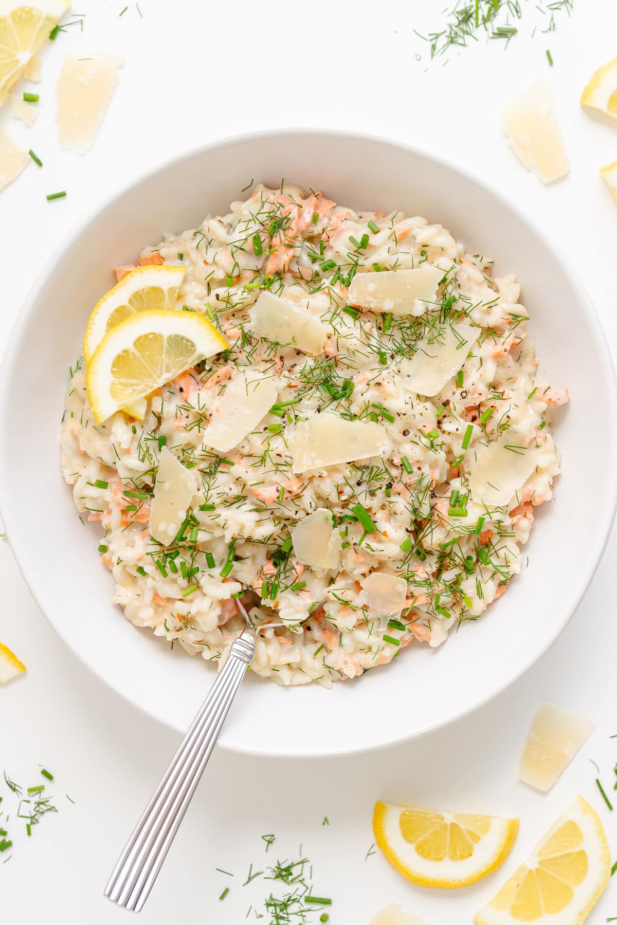 Salmon risotto in white bowl with spoon, garnished with lemon slices, parmesan shavings, fresh dill and chives.