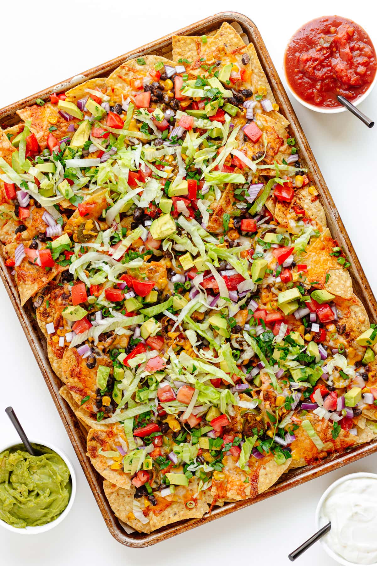 Vegetarian sheet pan nachos with small bowls of salsa, guacamole and sour cream beside it.