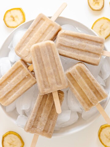 Banana popsicles sitting on a white platter of ice with banana slices scattered around it.