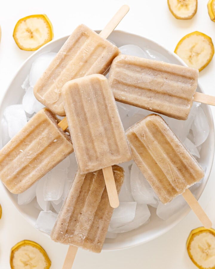 Banana popsicles sitting on a white platter of ice with banana slices scattered around it.