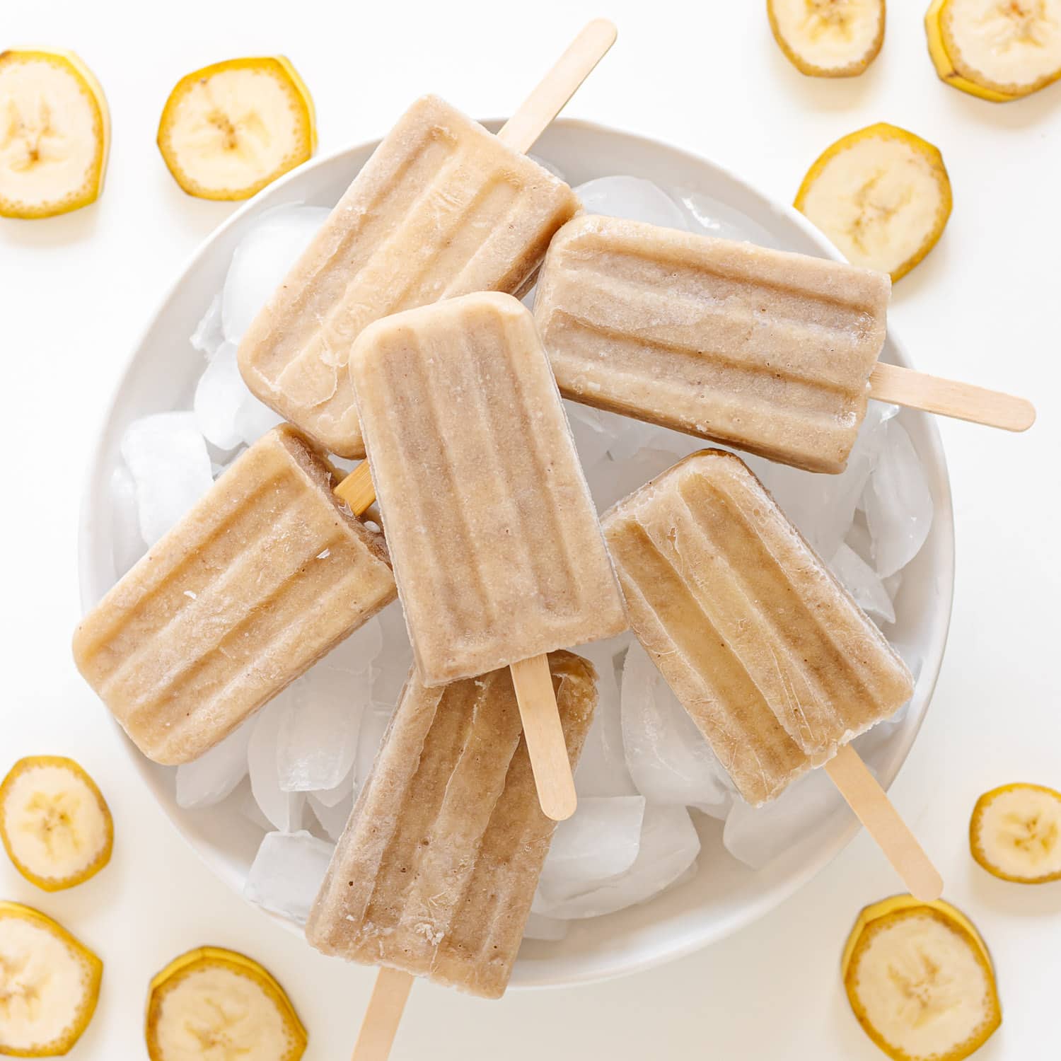 Banana popsicles sitting on a white platter of ice with banana slices scattered around it.