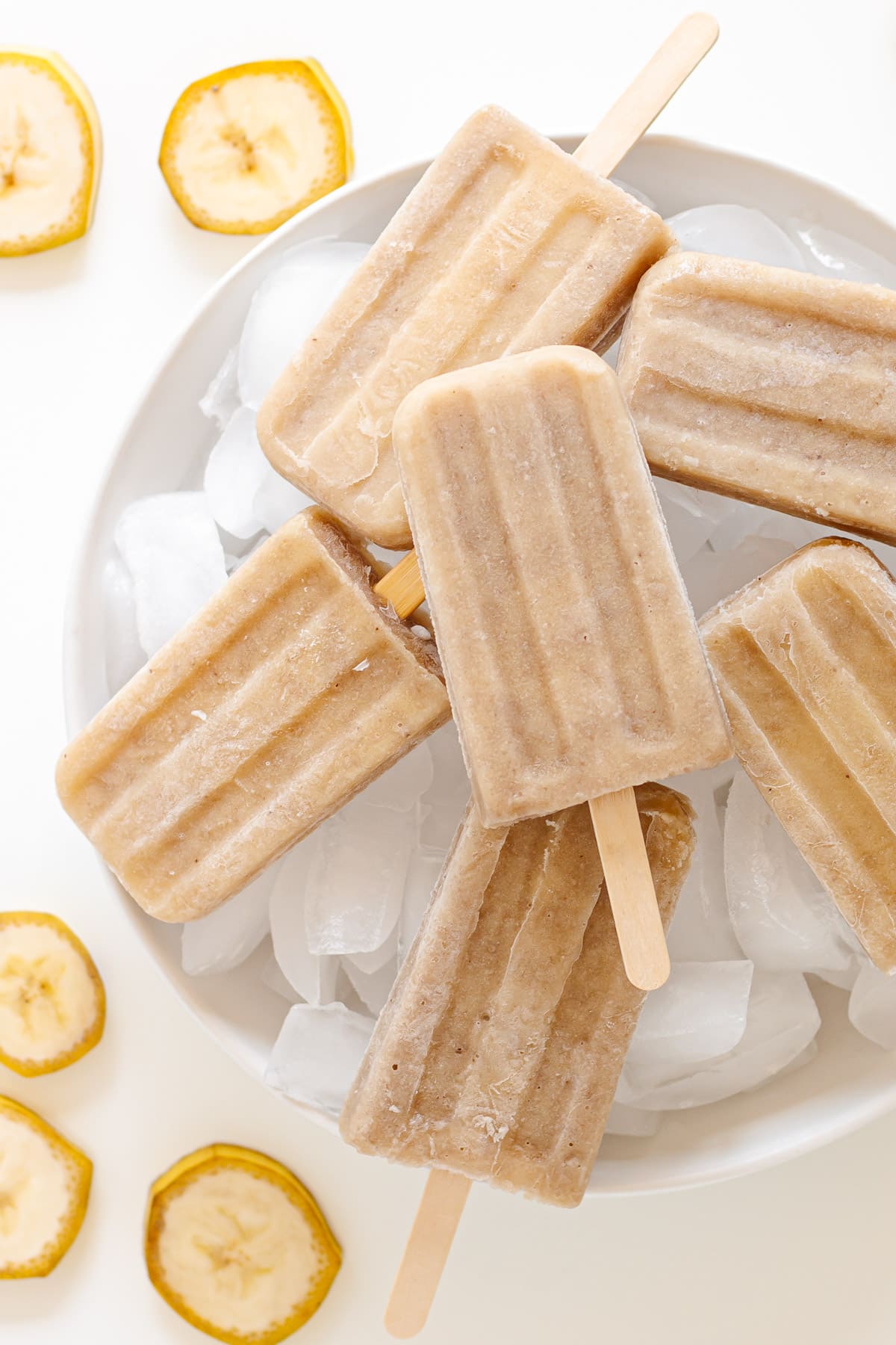 Banana popsicles sitting on a round white platter of ice cubes with slices of banana beside it.