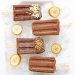 Chocolate banana popsicles sitting on ice with banana slices scattered around them.