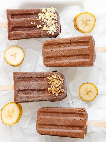 Chocolate banana popsicles sitting on ice with banana slices scattered around them.