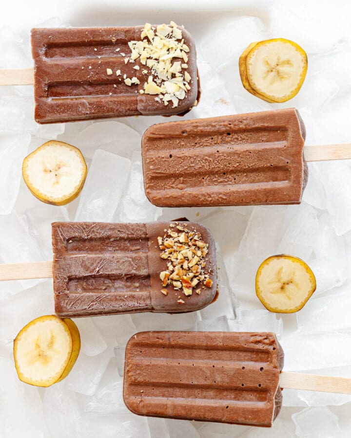 Chocolate banana popsicles sitting on ice with banana slices scattered around them.