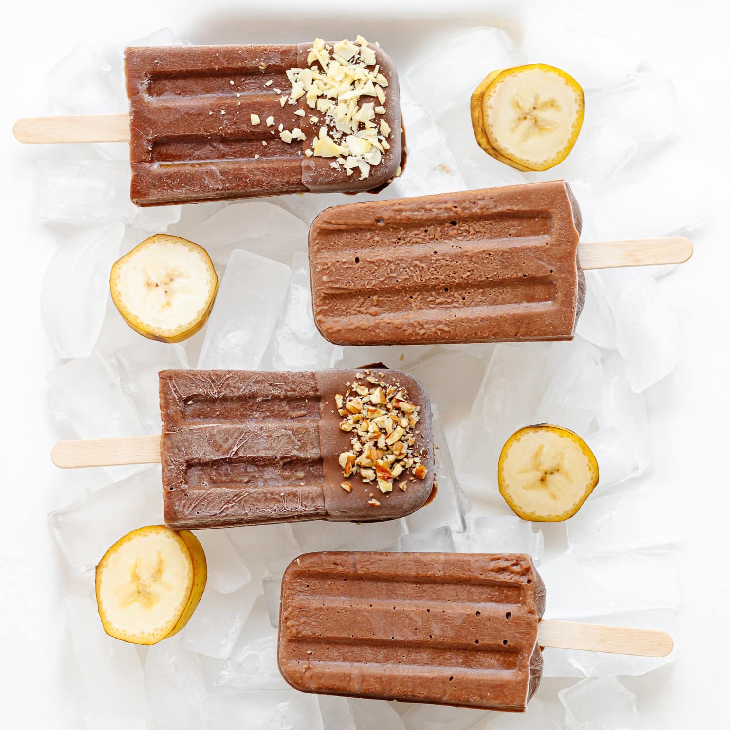 Chocolate banana popsicles sitting on ice with banana slices scattered around them.