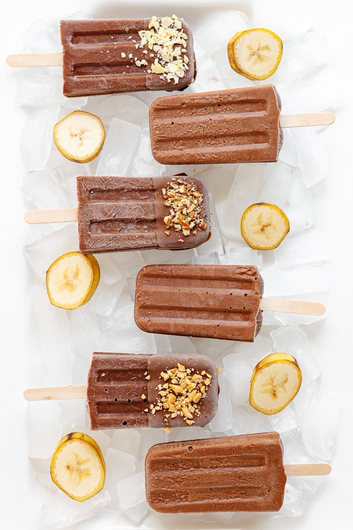 Chocolate banana popsicles sitting on a white platter of ice with banana slices.