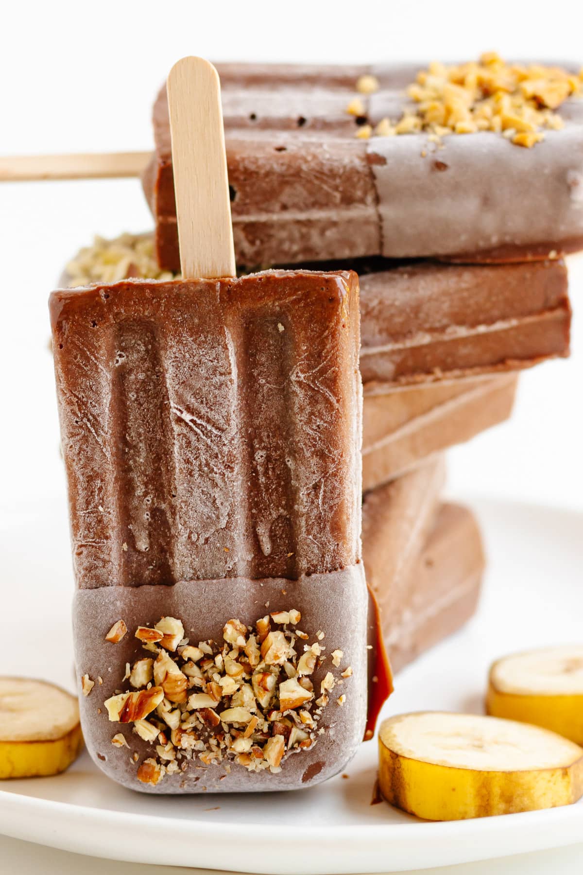 Chocolate banana popsicle dipped in chocolate and chopped nuts leaning against a pile of popsicles.