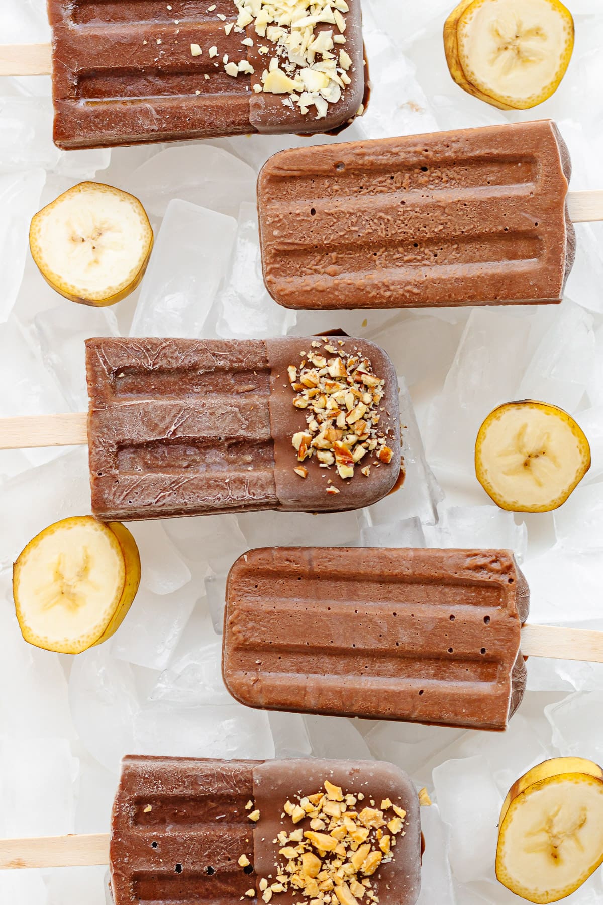 Chocolate banana popsicles sitting on ice with banana slices scattered around them.