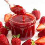 Spoonful of strawberry compote being lifted out of a glass jar surrounded by fresh strawberries.