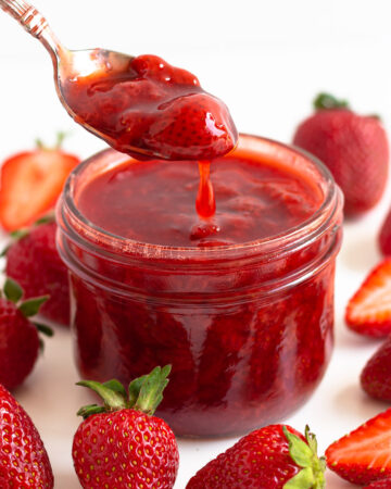 Spoonful of strawberry compote being lifted out of a glass jar surrounded by fresh strawberries.