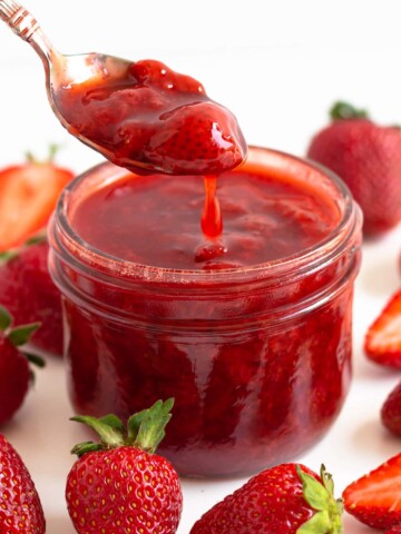 Spoonful of strawberry compote being lifted out of a glass jar surrounded by fresh strawberries.