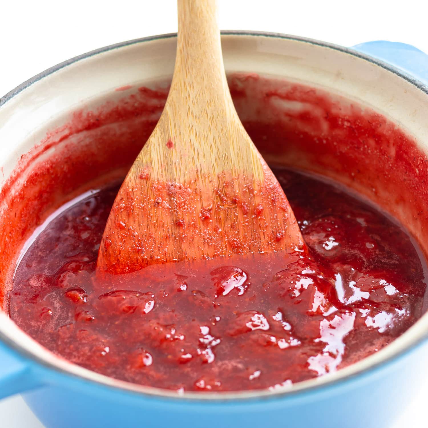 Wooden spoon stirring strawberry compote in saucepan.