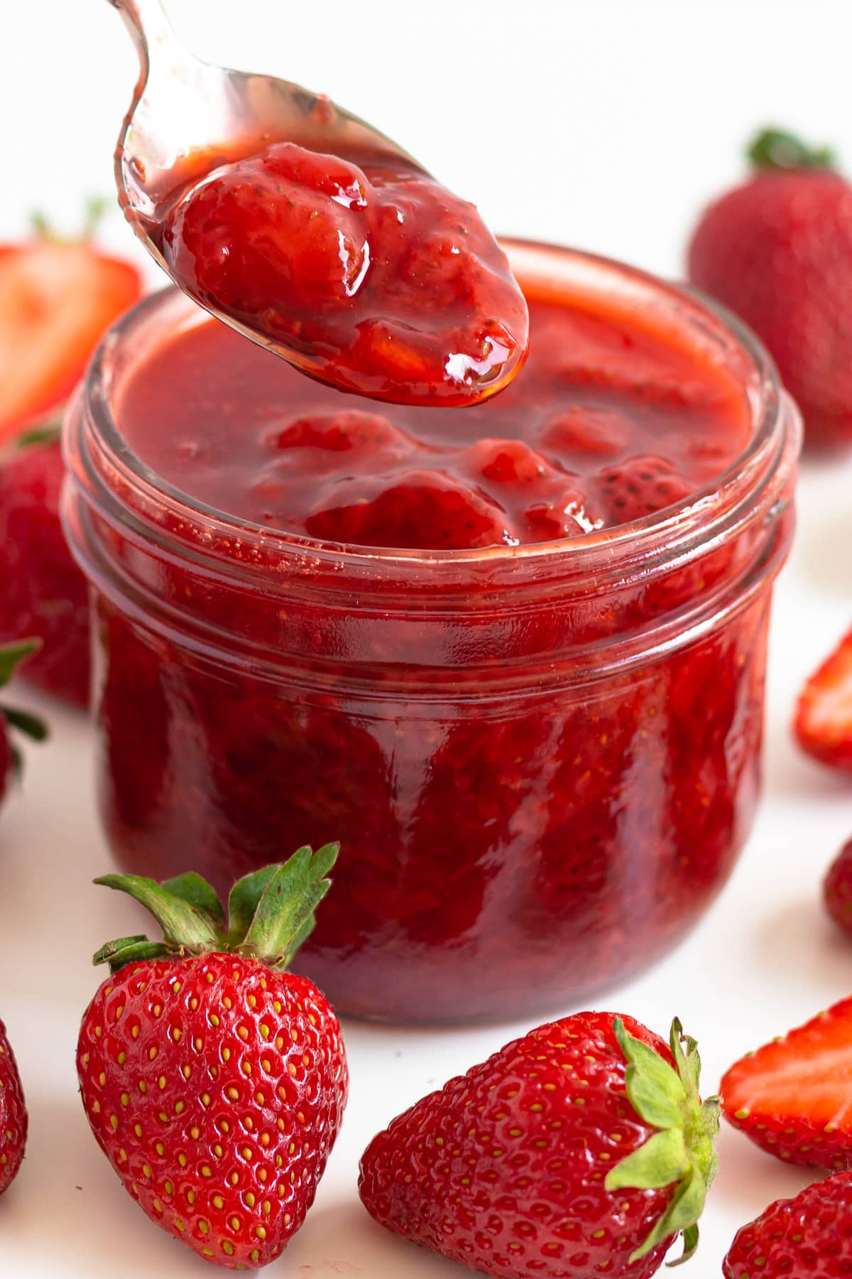 Jar of strawberry compote surrounded by fresh strawberries and spoon scooping some out.