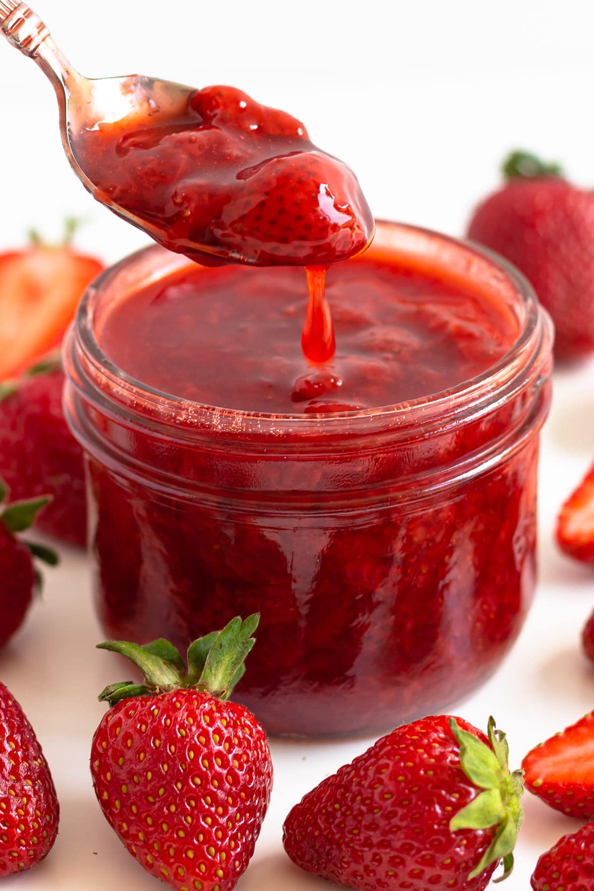 Strawberry compote dripping off a spoon into a jar surrounded by fresh strawberries.