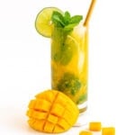 A mango mojito garnished with fresh mint and slice of lime with cut mango sitting beside glass.