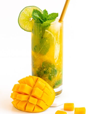 A mango mojito garnished with fresh mint and slice of lime with cut mango sitting beside glass.