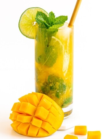 A mango mojito garnished with fresh mint and slice of lime with cut mango sitting beside glass.